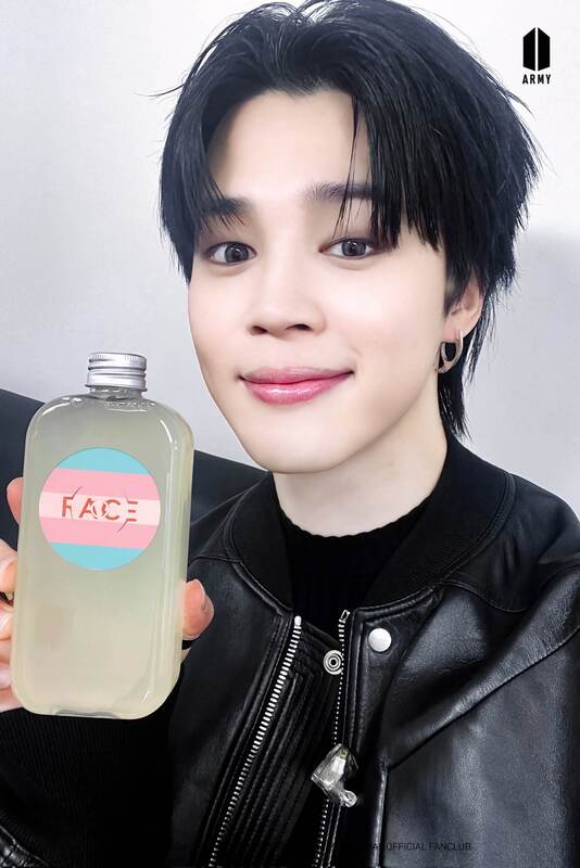jimin holding up a bottle with a circular face sticker on it that is trans flag colored