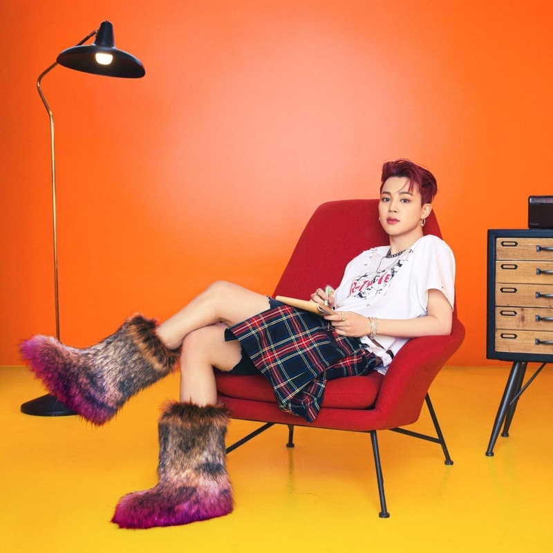 jimin from butter concept photos sitting in an orange toned room on a red chair in a white shirt and dark plaid kilt with his legs crossed toward the side of the chair, showcasing him wearing big fuzzy blurred asexual flag boots