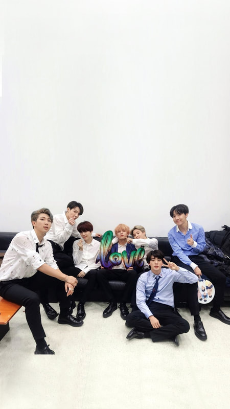 BTS on a couch from Taehyung's surprise birthday party in 2019 where Taehyung is sitting in the middleo f the members holding a balloon that says love. The balloon is blurred ocean gay flag colored.